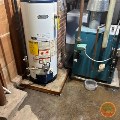A traditional Whirlpool tank-style water heater installed in a basement, standing on a wooden platform. The heater is vented through metal ducting that connects to a chimney. Next to it is a green boiler unit with connected piping and a small plastic bowl on the floor, possibly to catch drips. The concrete floor shows signs of water staining or leakage, and a brick wall is visible behind the setup.